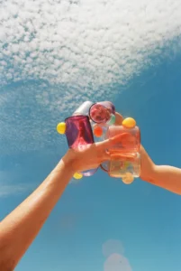 Two hands toasting with Bubble Cups in hand-blown glass, one filled with a pink drink, against a blue sky with clouds, by Sticky Glass